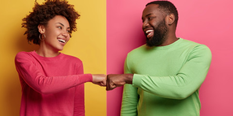 positive-dark-skinned-young-woman-and-man-bump-fists-agree-to-be-one-team-look-happily-at-each-other-celebrates-completed-task-wear-pink-and-green-clothes-pose-indoor-have-successful-deal